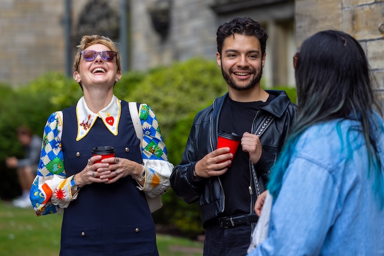 Three people chat outdoors while holding takeaway coffee cups; two are smiling and laughing.